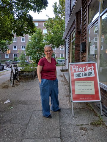 Wiebke Fuchs (Die Linke) vor dem Büro der Partei Die Linke. Rotes Oberteil, blaue Jeans, freundliches Gesicht.