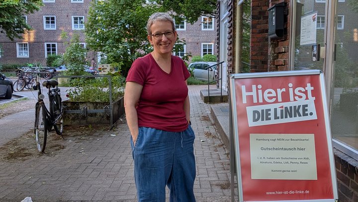 Wiebke Fuchs (Die Linke) vor dem Büro der Partei Die Linke. Rotes Oberteil, blaue Jeans, freundliches Gesicht.