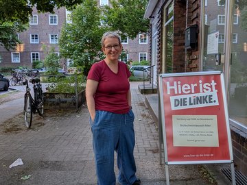 Wiebke Fuchs (Die Linke) vor dem Büro der Partei Die Linke. Rotes Oberteil, blaue Jeans, freundliches Gesicht.